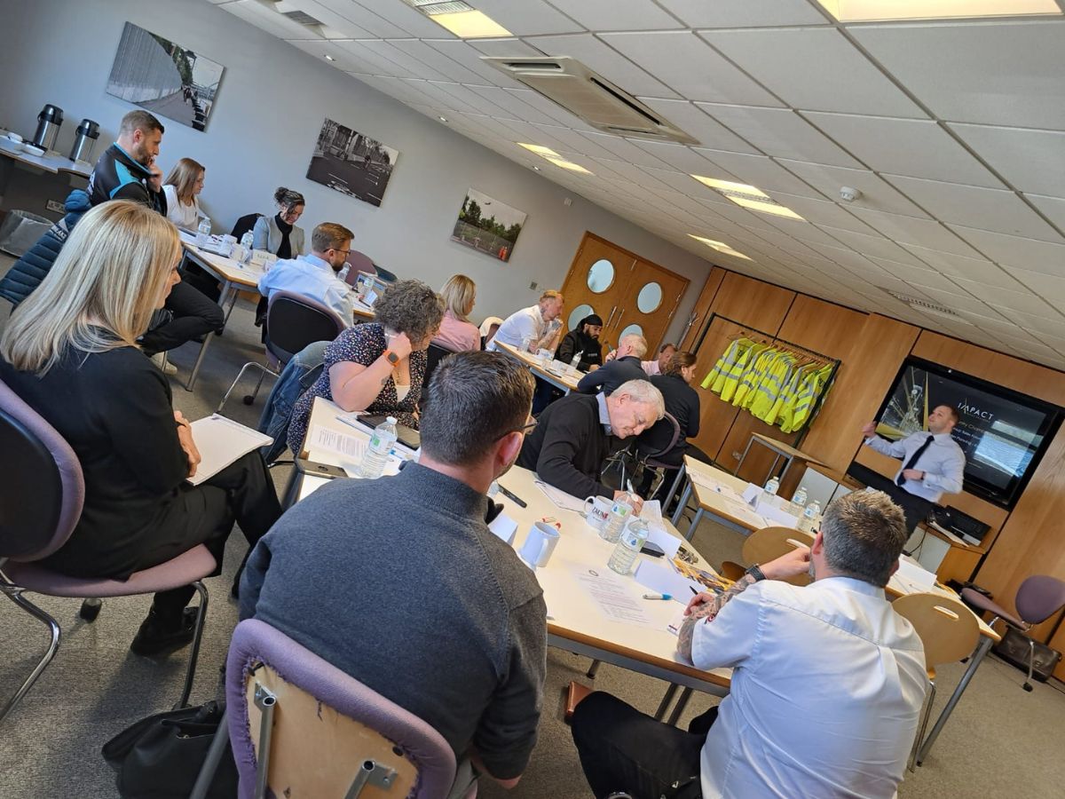 An image of a peer-to-peer group meeting, with Platinum Group members sat around 3 tables with drinks and notebooks. Adam Wootton from Impact Sales Coaching presents in front a projector.